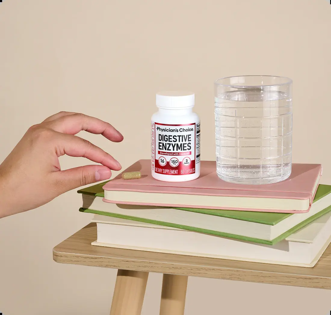 A hand reaching for a Physician's Choice Digestive Enzymes bottle and a capsule resting on a stack of books, next to a glass of water