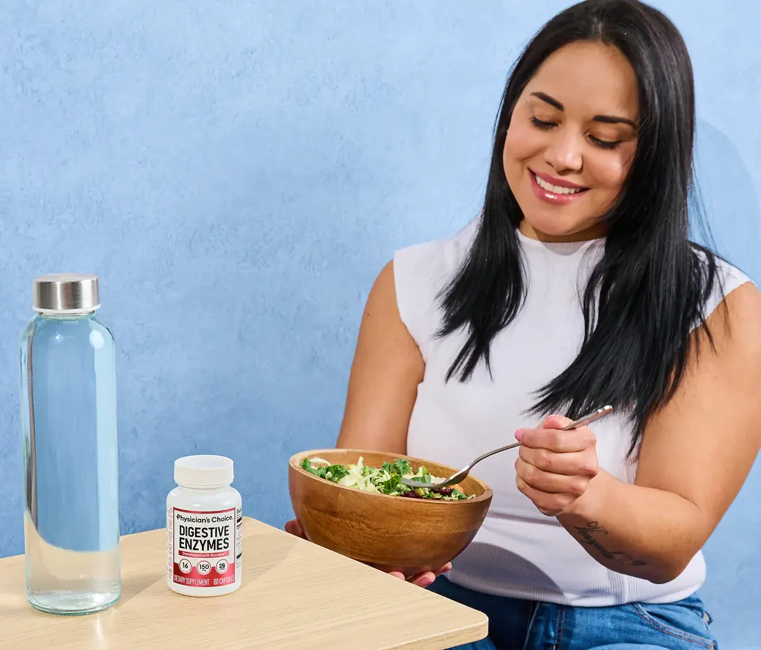 A woman smiling while eating a salad, with a glass water bottle and a Physician's Choice Digestive Enzymes bottle on the table beside her